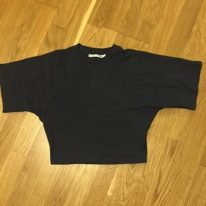 Svart croppad topp från 157 - En svart croppad topp från 157 med rund halsringning och vida korta ärmar. Enkel och stilren design som passar perfekt till jeans eller kjol. Tillverkad i mjuk bomull för en skön känsla hela dagen.