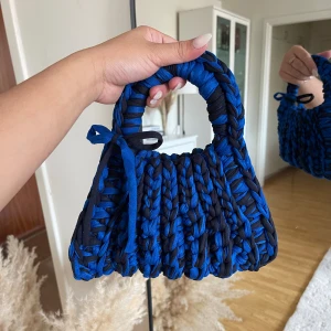 Blå och svart stickad handväska - Unik handväska i blått och svart, stickad med grova maskor och med en söt knytdetalj på sidan. Väskan har rundade handtag och en cool, chunky look som verkligen sticker ut. Perfekt accessoar för att lyfta din outfit.