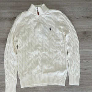 Vit kabelstickad tröja Polo Ralph Lauren - Kabelstickad vit tröja från Polo Ralph Lauren med halv dragkedja och hög krage. Klassisk design med broderad logga på bröstet och ribbade muddar. Perfekt för dig som gillar stilren och tidlös look.