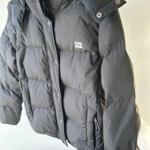 Svart dunjacka från Levi's med huva - Svart dunjacka från Levi's med vattenavvisande yta och varm stoppning. Jackan har huva, dragkedja och tryckknappar framtill samt praktiska fickor med dragkedja. Klassisk pufferstil med quiltade paneler och diskret logga på bröstet.