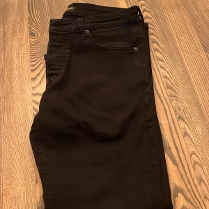 Svarta jeans från Jack & Jones - Svarta jeans från Jack & Jones med klassisk femficksdesign och diskret bakficka. Snygg, enkel look som passar till det mesta. Jeansen har smal passform och är tillverkade i ett stretchigt bomullsmaterial för extra komfort.