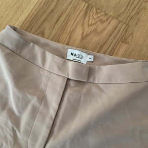 Beige kostymbyxor med slits från NA-KD, stl 34 - Snygga beige kostymbyxor från NA-KD med hög midja och raka ben. Byxorna har diskreta fickor bak och slits vid benslut. Perfekta för en clean och stilren look. Materialet känns mjukt och har en lätt stretch för extra komfort.