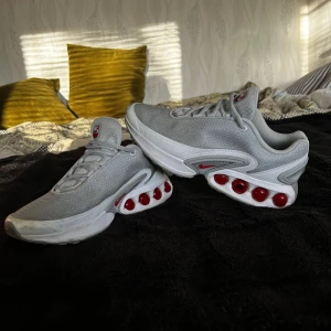 Nike Air Max DN grå sneakers - Nike Air Max DN sneakers i grått med röda detaljer och futuristisk design. Ovandel i mesh för bra andningsförmåga, synliga röda Air Max-kapslar i sulan och klassisk Nike-logga på sidan. Skorna har snörning och platt sula för en sportig look.