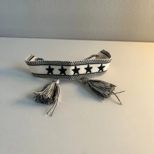 Svartvitt armband med stjärnor och tofsar - Snyggt armband i svart och vitt med broderade svarta stjärnor längs bandet. Armbandet har två dekorativa tofsar i grått och vitt som ger en cool och unik touch. Perfekt för dig som vill sticka ut med detaljer.