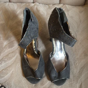 Svarta pumps med nitar och öppen tå - Snygga svarta pumps med öppen tå och klack. Skorna har ett brett band över vristen dekorerat med små nitar och är gjorda i mocka. Insidan har en metallic finish som ger en cool kontrast. Perfekta för dig som vill sticka ut lite extra.