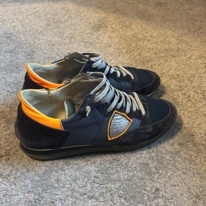 Philippe Model Trainers  - Snygga sneakers från Philippe Model i mörkblå mocka och skinn med grå meshdetaljer. Skorna har en orange hälkappa och silverfärgad sköld på sidan. Grå snörning och platt sula. Perfekta för dig som gillar unika detaljer och streetstyle. En liten defekt på andra bilden samt välanvända därav priset. Kontakta för frågor