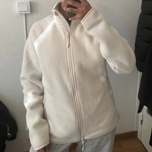 Vit fleecejacka med dragkedja - Mjuk och mysig vit fleecejacka med hög krage och hel dragkedja framtill. Jackan har två sidofickor med dragkedja och en enkel, clean design. Perfekt för chill dagar och när du vill hålla dig varm. Passformen är normal och ärmarna är långa.