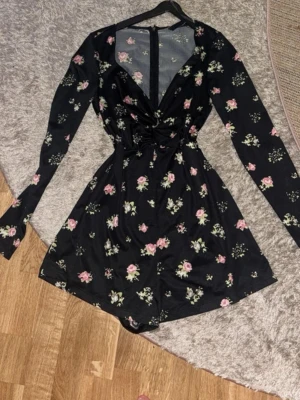 Svart blommig byxdress med v-ringning - Supersnygg svart byxdress med rosa och grönt blommönster. Långärmad modell med v-ringad omlott framtill och markerad midja. Perfekt för dig som vill ha en feminin och trendig look. Materialet känns mjukt och följsamt.