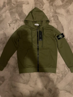 Olivgrön hoodie från Stone Island - Snygg olivgrön hoodie från Stone Island med dragkedja framtill, huva med dragsko och klassisk logopatch på vänster ärm. Tillverkad i mjukt bomullsmaterial och har känguruficka framtill. Perfekt för en avslappnad och trendig stil.