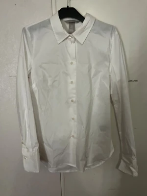 Vit klassisk skjortblus med krage - Stilren vit skjortblus med lång ärm och klassisk krage. Blusen har knäppning framtill med vita knappar och är tillverkad i ett slätt, vävt material. Perfekt för en clean och tidlös look. Aldrig använd. Med prislapp.