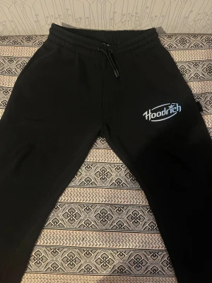 Svarta mjukisbyxor från Hoodrich - Hoodien finns också på min profil. Svarta mjukisbyxor från Hoodrich med vit och blå logga på ena benet. Byxorna har snörning i midjan och är gjorda i mjukt bomullsmaterial. Perfekta för chill eller streetwear-stil.