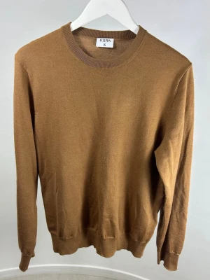 Filippa K Stickad Tröja - En stilren brungul stickad tröja från Filippa K med rund hals och långa ärmar. Modellen är 175 cm och väger 70 kg. Tröjan har ribbade muddar vid ärmslut och nederkant, vilket ger en snygg passform. Perfekt för lager-på-lager och enkel att matcha med jeans eller chinos.