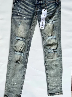Distressed jeans från Purple, blå - Snygga blå jeans från Purple med slitna detaljer och hål på benen. Jeansen har en tvättad look med ljusa partier och vita färgstänk för en trendig vibe. Klassisk femficksmodell och normal passform. Perfekta för dig som gillar streetstyle och edgy mode.