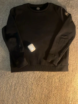 Svart sweatshirt från Colmar - Svart sweatshirt från Colmar Originals med rund halsringning och ribbade muddar. Klassisk och stilren design med Colmar-logga på ärmen. Perfekt för en avslappnad och trendig look.