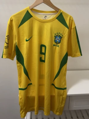Brasilien Ronaldo 9 matchtröja Nike - Gul Brasilien fotbollströja från Nike med gröna detaljer, nummer 9 och namnet Ronaldo på ryggen. Tröjan har korta ärmar och officiellt landslagsmärke på bröstet. Perfekt för fotbollsfans som vill sticka ut på läktaren eller planen.