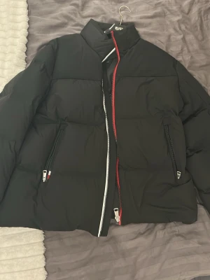 Svart pufferjacka från Tommy Hilfiger - Svart pufferjacka från Tommy Hilfiger med hög krage och två dragkedjefickor framtill. Jackan har en unik dragkedja i vitt och rött samt diskret logga broderad på kragen. Perfekt för kyliga dagar. Använd max en månad. Köpt för 1800kr på rea 