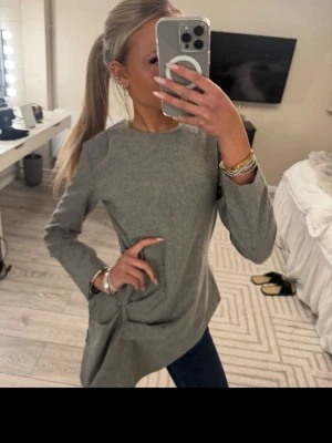 Grå långärmad blus med asymmetrisk fåll - Stilren grå blus med lång ärm och rund halsringning. Blusen har en snygg asymmetrisk fåll och är tillverkad i ett mjukt, vävt tyg som ger en clean look. Perfekt att matcha med jeans eller kjol för en trendig outfit.