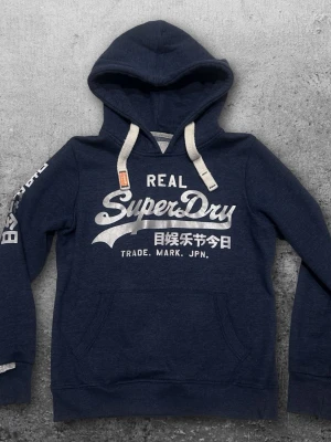 Superdry hoodie XS - En lite schysstare hoodie från Superdry i färgen mörkblå med silvrigt tryck. Storlek XS även fast det står S. Mycket bra skick. 😀