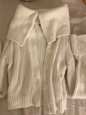 Vit stickad kofta med stor krage - Säljer en vit stickad kofta med stora knappar och en bred, nedvikt krage. Koftan har ribbstickad struktur och långa ärmar. 