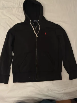 Svart hoodie med dragkedja från Polo Ralph Lauren - Svart hoodie med dragkedja från Polo Ralph Lauren. Klassisk design med vit dragsko i huvan, känguruficka och röd broderad logga på bröstet. Mjuk bomullskänsla och ribbade muddar vid ärmslut och nederkant. Byxor från samma set finns kolla profil.
