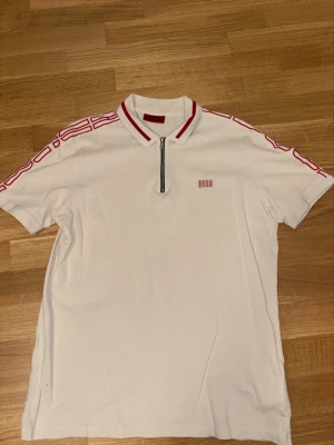 Vit polotopp från Hugo med dragkedja - Vit polotopp från Hugo med röda detaljer på krage och axlar samt röd logga på bröstet. Toppen har korta ärmar och en snygg dragkedja framtill. Tillverkad i bomull för en skön känsla och sportig look.