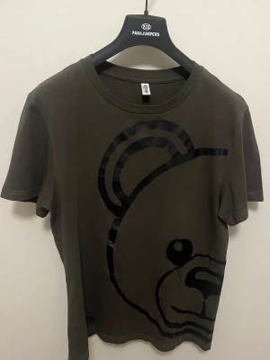 Olivgrön t-shirt med björntryck - Cool olivgrön t-shirt från Moschino med ett stort svart björntryck som går från framsidan till baksidan. T-shirten har rund halsringning och korta ärmar. Perfekt för dig som gillar statement-plagg och streetstyle.