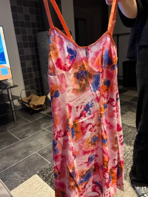 Färgglatt nattlinne med blommigt mönster - Säljer ett nattlinne med tunna axelband och färgstarkt blommigt mönster i rosa, orange, blått och vitt. Materialet känns lätt och svalt, perfekt för att chilla hemma. Snygg passform med markerad byst och mjuk känsla. Står ej varumärket. Aldrig använd 
