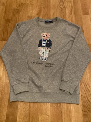 Grå sweatshirt Polo Bear Ralph Lauren - Grå sweatshirt från Polo Ralph Lauren med ikoniskt Polo Bear-tryck framtill. Rund halsring och avslappnad passform. Perfekt för en cool och avslappnad stil. OBS storleken på tröjan är M men den är liten till storleken så den passar S perfekt.      Priset går att diskutera!!