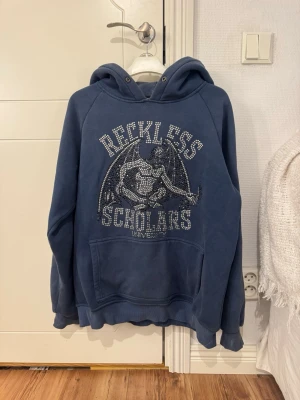 Blå hoodie Reckless Scholars - Mörkblå hoodie med tryck och strasstenar framtill från Reckless Scholars. Stor huva och känguruficka. Snyggt motiv med demon och text. Perfekt för dig som gillar streetwear och vill sticka ut.