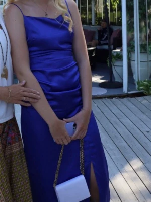 Lång blå satinklänning med slits - Elegant lång klänning i glansigt blått satintyg med smala axelband och hög slits framtill. Figurnära passform och enkel, stilren design som ger en lyxig känsla. Perfekt för speciella tillfällen.