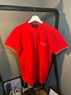 Röd kortärmad piké från Sail Racing - Snygg röd pikétröja från Sail Racing med klassisk krage och knappar framtill. Diskret logga på bröstet och vit detaljsöm vid ärmslut. Perfekt till jeans eller shorts för en sportig look.