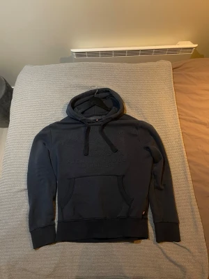 Mörkblå hoodie från Superdry - Snygg mörkblå hoodie från Superdry med broderad logga på bröstet och klassisk känguruficka. Hoodien har dragsko i huvan och ribbade muddar vid ärmar och nederkant. Perfekt för en avslappnad och trendig stil.