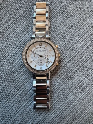 Michael kors klocka  - Michael Kors Parker MK5626 . Vit/Gulguldtonat stål Ø39 mm.  Michael Kors damklocka med guldtonad metallboett och armband. Urtavlan är vit med guldfärgade visare och index, inklusive tre mindre urtavlor för kronograffunktioner. Klockan har kristalldetaljer runt boetten.Armbandet är tillverkat i rostfritt stål. klockan behöver NYA batterie. Min handled är 16cm oxh kan ha klockan på mig , vet ej storlek på länken.