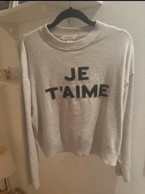 Grå sweatshirt med tryck 'JE T'AIME' - Trendig grå sweatshirt med svart text 'JE T'AIME' framtill. Mjuk bomullskänsla, rund halsringning och långa ärmar. Perfekt till jeans eller kjol för en avslappnad look.