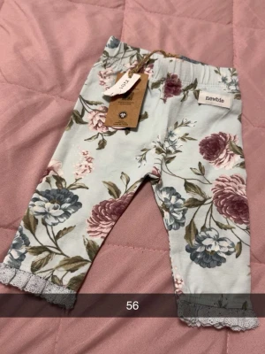 Blommiga byxor från Newbie - Supersöta blommiga byxor från Newbie i ljusblått med spetsdetaljer vid bensluten. Byxorna har elastisk midja och ett romantiskt blommönster i blått, lila och grönt. Perfekta för dig som gillar gulliga och bekväma plagg till de minsta.