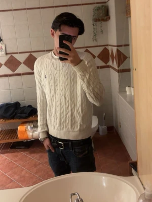 Vit kabelstickad tröja från Ralph Lauren - Vit kabelstickad tröja från Ralph Lauren med rund halsringning och diskret broderad logga på bröstet. Klassisk och stilren design med ribbade muddar vid ärmslut och nederkant. Perfekt för en snygg och avslappnad look.