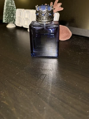 K by Dolce & Gabbana parfym - En lyxig parfym från Dolce & Gabbana i en mörkblå glasflaska med en unik kronformad kork dekorerad med blå stenar. Flaskan har en stilren, fyrkantig design och utstrålar exklusivitet och elegans. Runt 35-40 ml kvar