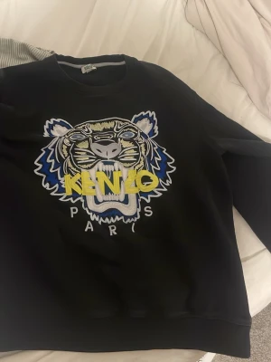 Svart Kenzo hoodie med tigertryck - Svart hoodie från Kenzo med ikoniskt broderat tigerhuvud i blått, vitt och gult framtill. Stora bokstäver 'KENZO PARIS' över motivet. Klassisk passform och rund halsringning. Perfekt för dig som vill sticka ut med en streetstyle-look.