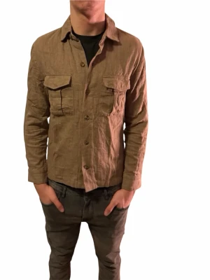 Beige overshirt i lin från Riley - Säljer en snygg beige overshirt från Riley i 100% linne. Jackan har två stora bröstfickor med lock, knäppning framtill och klassisk krage. Perfekt för lager-på-lager och ger en chill vibe till outfiten.