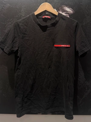 Svart t-shirt från Prada - Snygg svart t-shirt från Prada med klassisk passform och korta ärmar. På bröstet finns en röd och vit logga som ger en stilren touch. Perfekt för dig som gillar exklusiva märken och enkel design.
