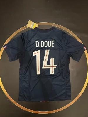PSG matchtröja D. Doué #14 - Mörkblå Paris Saint-Germain matchtröja från Nike med D. Doué och nummer 14 på ryggen. Tröjan har korta ärmar, klubbmärke på bröstet och Qatar Airways som sponsor framtill. Snyggt rödblått mönster längs mitten. Perfekt för fotbollsfans!
