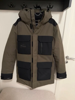 Mörkgrön glacier bay parka Sail Racing - Stilren parkajacka från Sail Racing i olivgrönt med svarta detaljer. Jackan har fyra stora fickor framtill, huva och dragkedja. Perfekt för kalla dagar med sin långa modell och praktiska design. OBS! Använd endast 2 gånger då jag köpt en ny jacka 