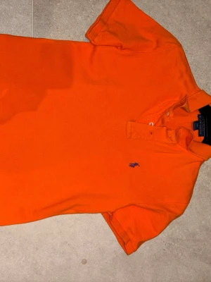 Orange pikétröja från Ralph Lauren - Klassisk orange pikétröja från Ralph Lauren med broderad logga på bröstet. Tröjan har krage, korta ärmar och knäppning framtill. Perfekt för dig som gillar färg och stilren design.