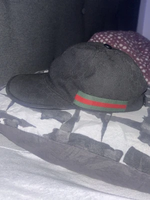 Svart keps från Gucci med röd/grön detalj - Svart keps från Gucci med klassisk röd och grön rand på sidan. Kepsen har böjd skärm och justerbar rem baktill. Materialet är tyg med diskret mönster i svart. Perfekt för dig som vill ha en stilren accessoar med lyxig känsla.