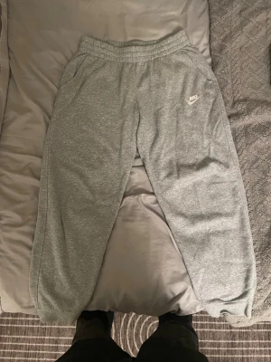 Grå mjukisbyxor från Nike - Klassiska grå mjukisbyxor från Nike med resår i midjan och vid bensluten. Byxorna har sidofickor och en liten vit Nike-logga på vänster lår. Perfekta för chill eller träning.