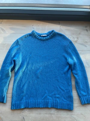 Blå stickad tröja från Zara strl L - En enkel och snygg blå stickad tröja från Zara med rund halsringning och ribbade muddar vid ärmslut och nederkant. Perfekt för lager på lager och passar till det mesta. Materialet ser mjukt och bekvämt ut.