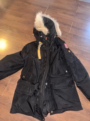 Svart Parajumpers jacka -  Parajumpers kodiak i storlek XL passar L utmärkt.  avtagbar huva och pälskant. Jackan har flera stora fickor, gul detalj vid halsen och midjan samt patch på ärmen. Perfekt för kalla dagar med sin långa passform och robusta dragkedja. kemtvättad o klar. fluffig päls.  inga riv eller risp märken. dock väger jackan en del så hängaren i jackan gått av ”se bild” 