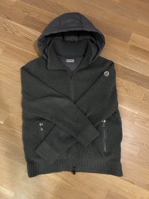 Grå Moncler pufferjacka med huva - Säljer en grå Moncler pufferjacka med stickade  ärmar och ribbade muddar.                                                      Den är helt nytt skick inga skador storlek L men jag skulle säga den passa bra för både M och L 