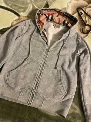 Grå zip hoodie från Burberry - Snygg grå hoodie med dragkedja från Burberry. Klassisk huva med det ikoniska rutmönstret på insidan och justerbara snören. Mjuk bomullskänsla, två stora fickor fram och ribbade muddar. Perfekt för en chill och stilren look. Pris kan diskuteras.