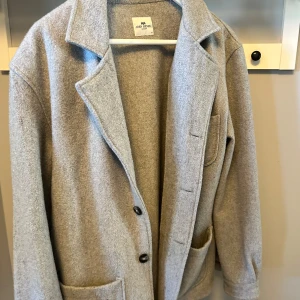 Grå kappa från John Henric - Snygg grå kappa från John Henric i storlek M. Jackan har klassisk krage, två stora framfickor och knäppning med svarta knappar. Tillverkad i mjuk ull som ger en stilren och clean look. Perfekt för dig som gillar minimalistisk stil.